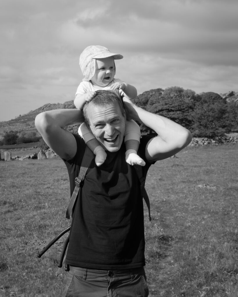 Ben Shears smiles at camera with daughter on shoulders, whilst walking on Dartmoor.